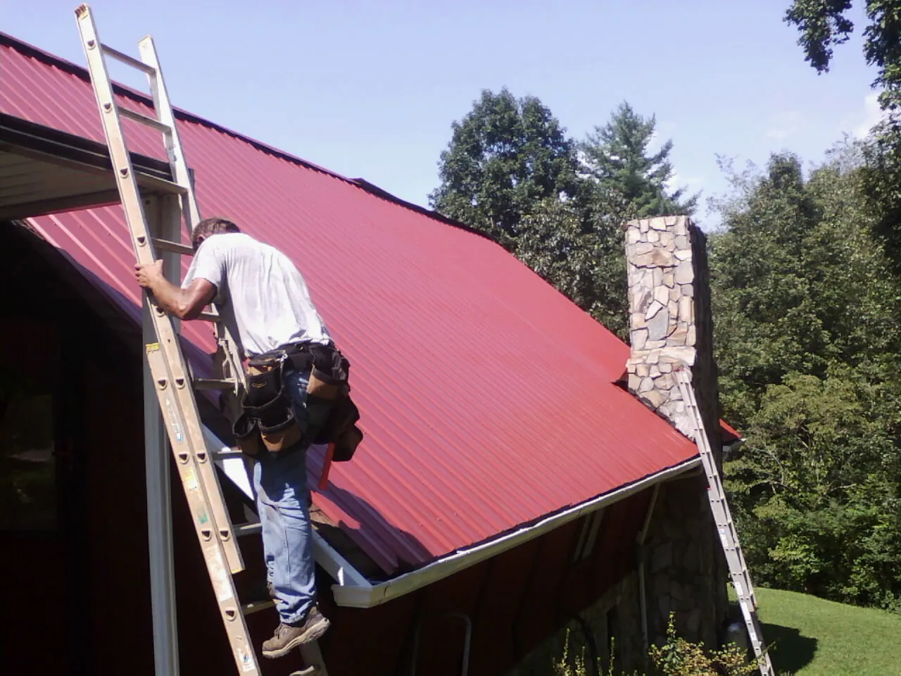 Roofer on a ladder beside a red metal roof and stone chimney — Soffit & Fascia Repair by Roofing Brothers in Fox Lake